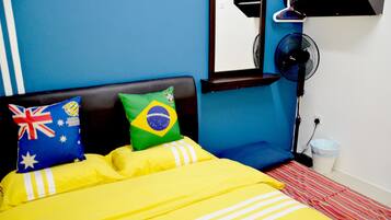 A bed with a yellow bedspread, pillows with flags, a standing fan, and a white trash bin.