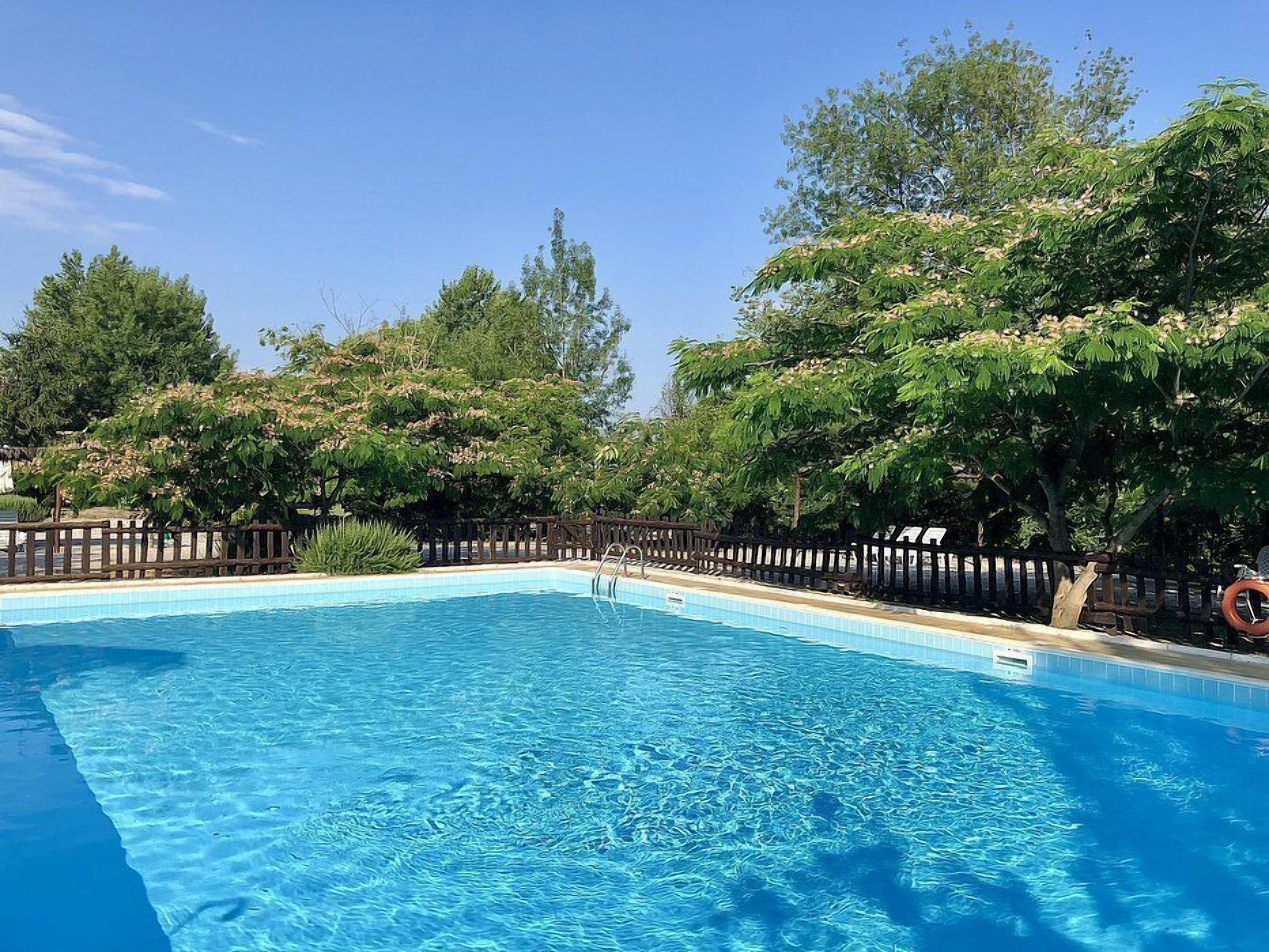 A rectangular swimming pool with clear blue water, surrounded by a wooden fence and lush green trees.