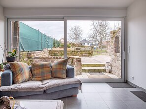 Un salón moderno con un sofá gris, cojines escoceses y vistas a un espacio exterior con un muro de piedra y una valla de malla verde.