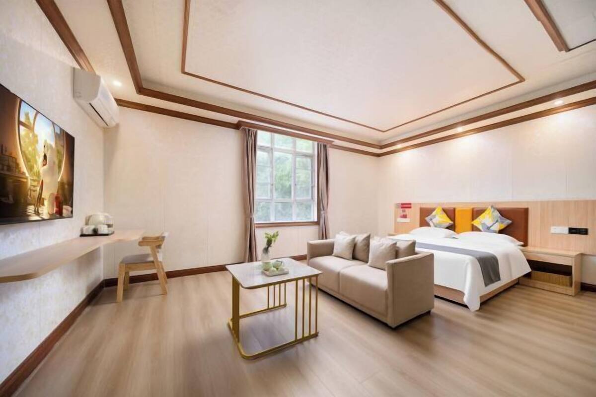 A hotel room with a bed, a sofa, a small table, a chair, a painting on the wall, and a window.
