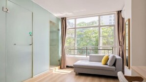 A modern room with a grey sofa, a large window with a view of greenery, and a wooden bench. - Shell Hotel (Haikou hospital Hainan University store) (Qiongshan)