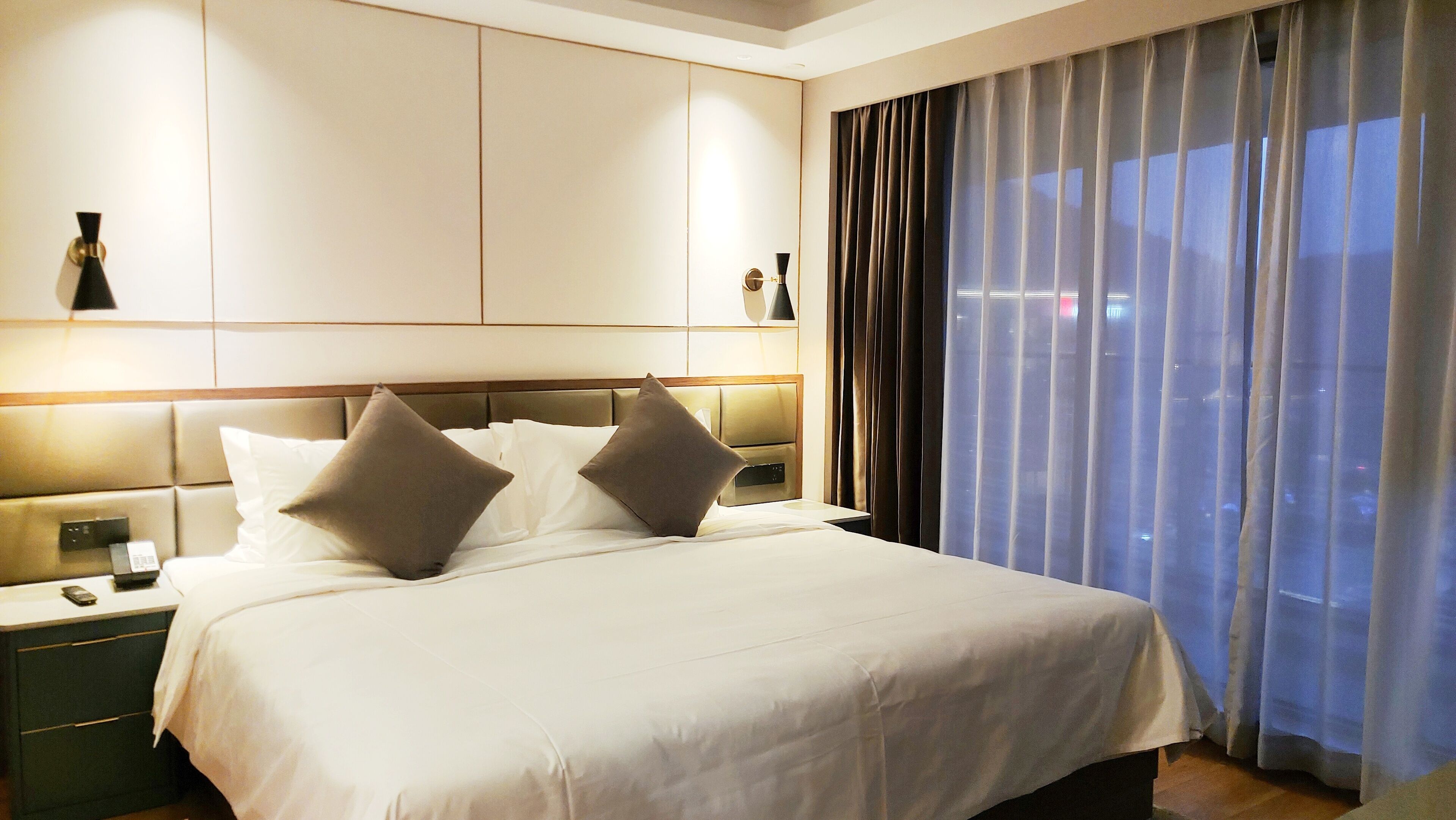 A hotel room with a large bed, a nightstand with a phone, and a view of the city through sheer curtains.