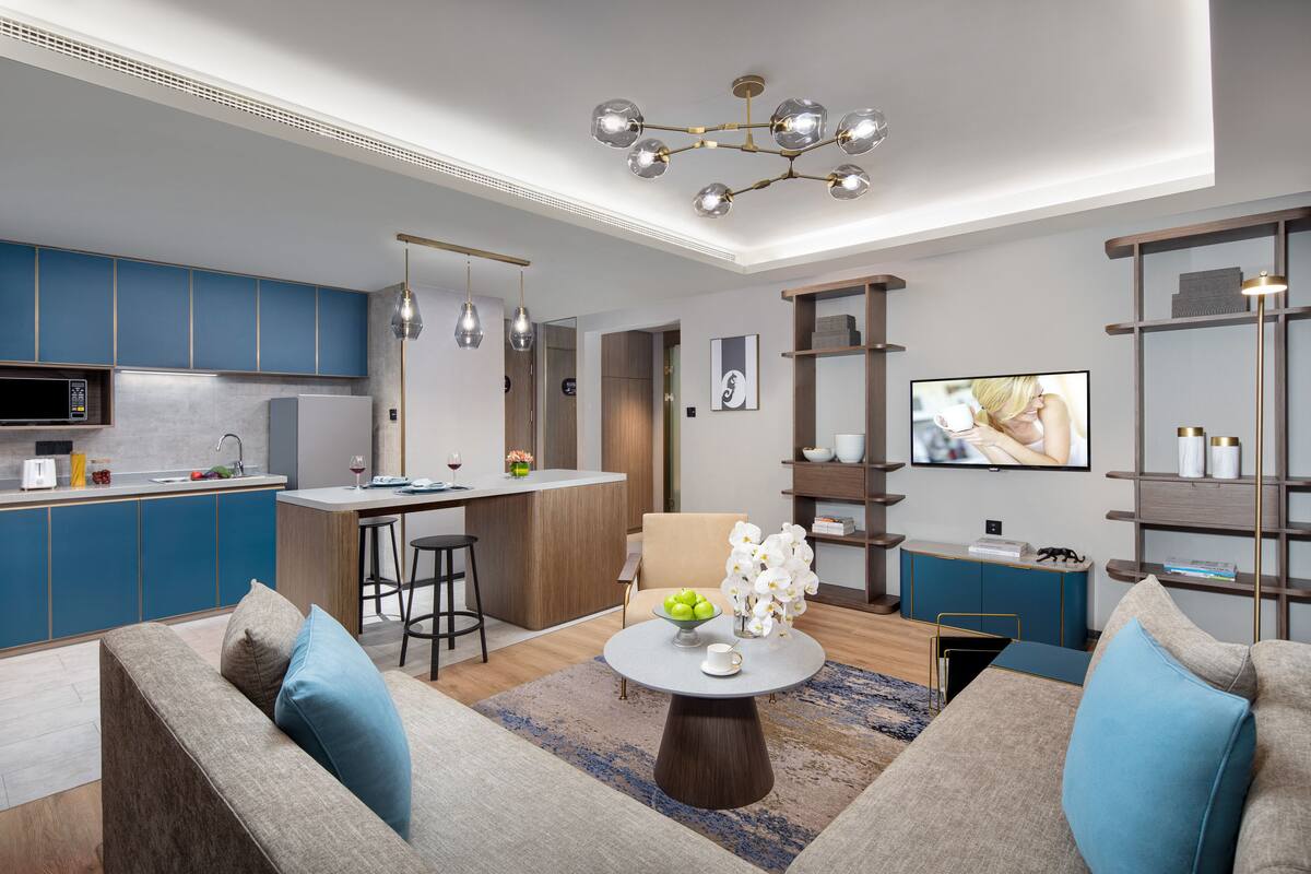 Una sala de estar moderna con un sofá, una mesa de centro y un televisor. Hay una zona de cocina con armarios azules y una mesa de comedor.
