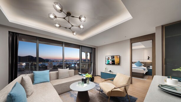 Une chambre d’hôtel moderne dotée d’une grande fenêtre donnant sur la ville, d’un canapé, d’un fauteuil, d’une petite table avec un vase et d’un lit avec un oreiller bleu.