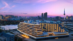 Un hôtel moderne à plusieurs étages, doté d’une terrasse sur le toit, et entouré d’un paysage urbain avec une tour imposante en arrière-plan.