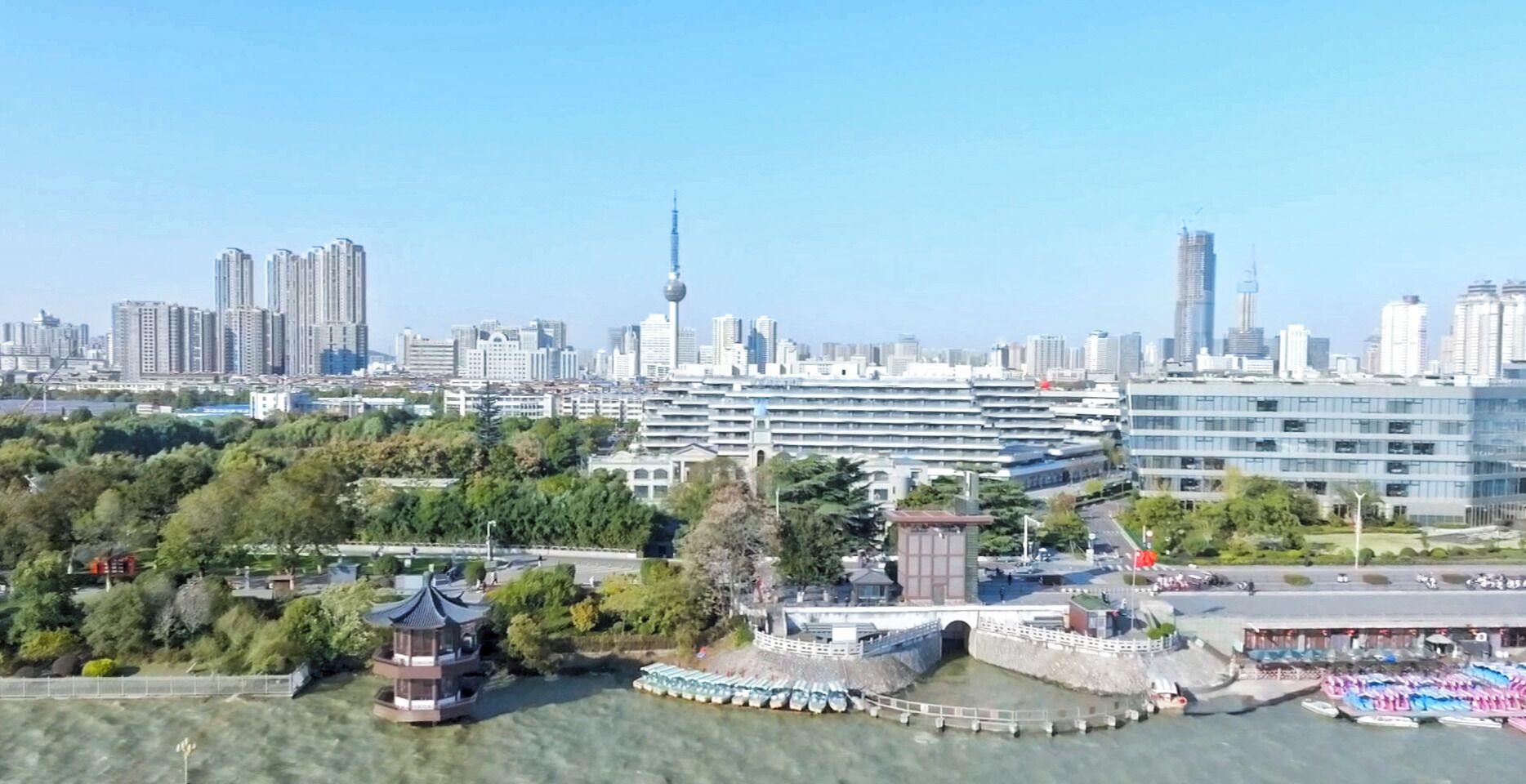 Panoramica urbana con moderni grattacieli, un'imponente torre e una zona sul lungomare con una struttura simile a un pagodae imbarcazioni.