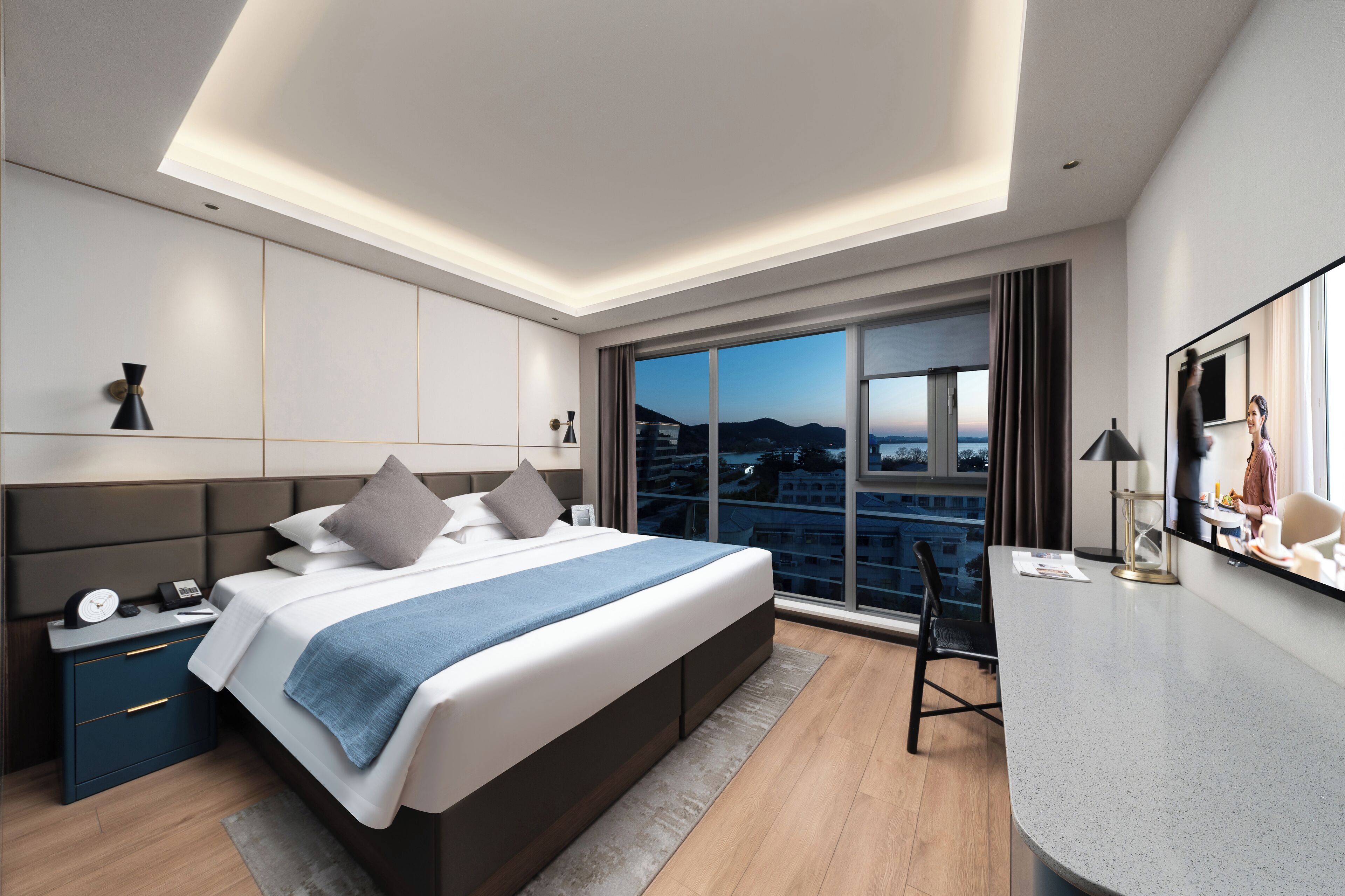 A modern hotel room with a large bed, bedside tables, a desk, and a large window offering a city view.