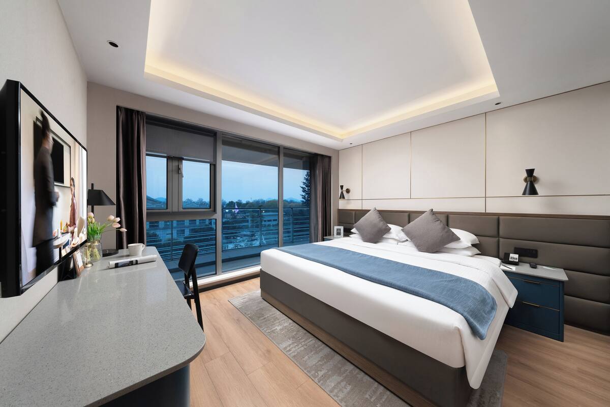 Habitación de hotel moderna con una cama grande, un escritorio y vistas de la ciudad a través de ventanas de piso a techo.