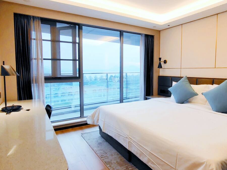 A hotel room with a large bed, a desk with a chair, a TV, and a view of the city through large windows.