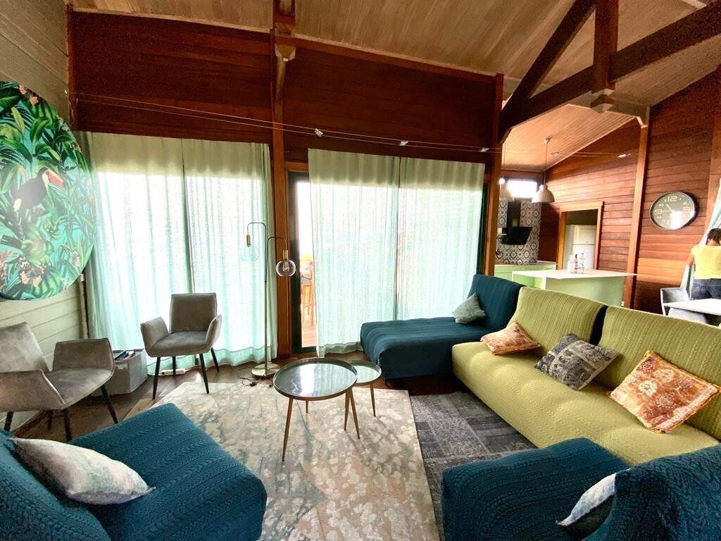 A cozy living room with a sofa, chairs, and a coffee table. There is a large window with curtains, a clock on the wall, and a plant on the left.