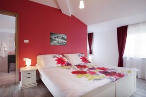 Une chambre avec un mur rouge, un lit recouvert d’une couette à motifs floraux, une table de chevet avec une lampe et un tableau accroché au mur.