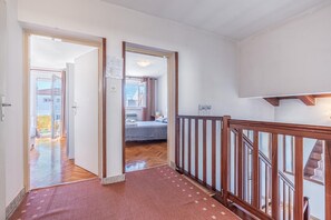 Een gang met houten leuning, vloerbedekking en uitzicht op een slaapkamer met een bed en een raam.