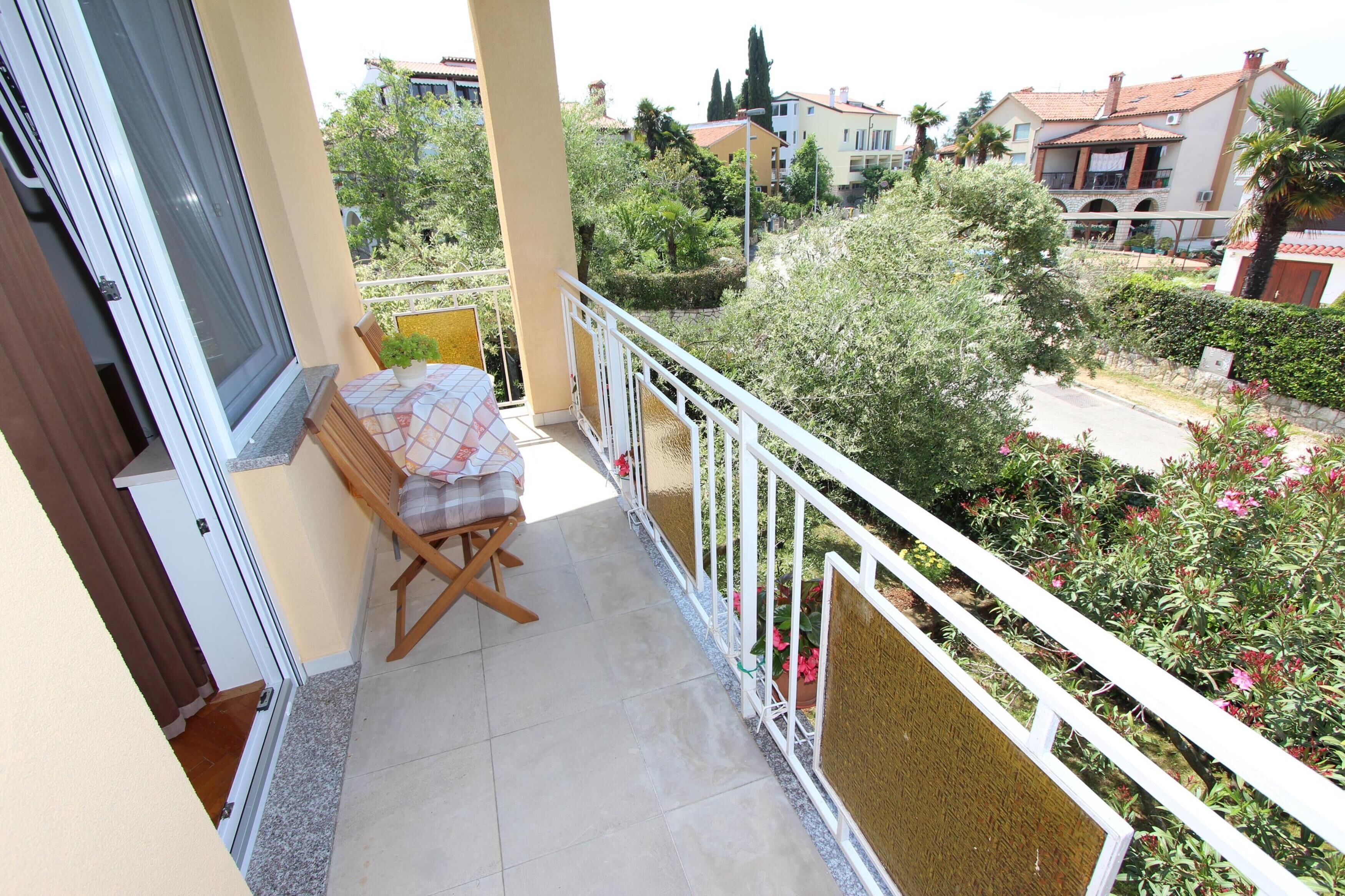 A balcony with a chair and table, overlooking a garden and neighboring buildings.