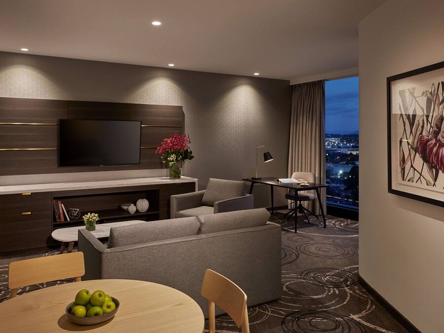 Habitación de hotel moderna con televisión de pantalla plana, sofá, mesa de comedor con manzanas, pequeño escritorio con lámpara y vista a la ciudad a través de la ventana.