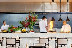 A modern restaurant with a central counter, chefs preparing food, and dining tables with chairs.