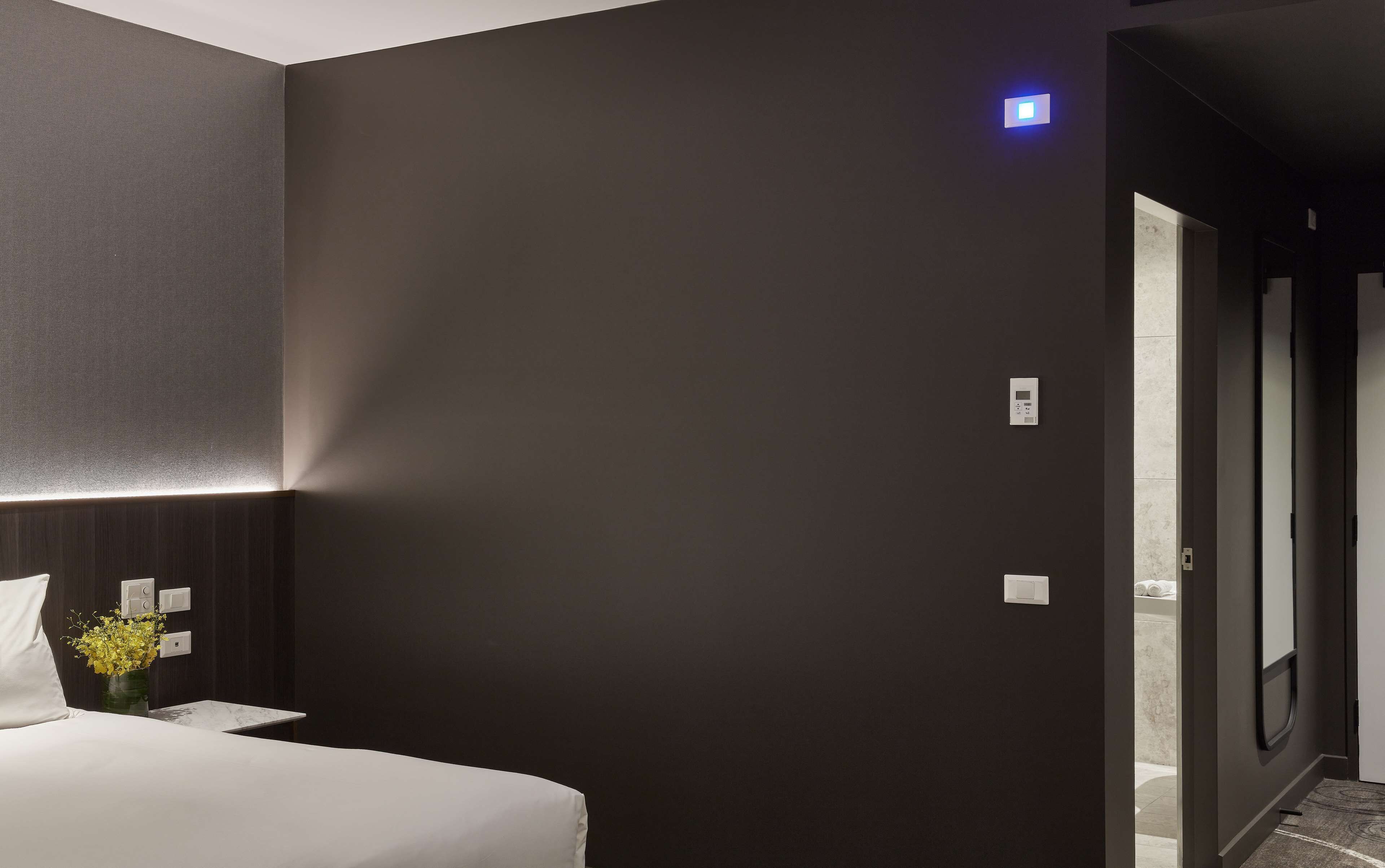 Quarto de hotel moderno com parede escura, cama com roupa de cama branca, mesa de cabeceira com um vaso de flores amarelas e uma porta visível que dá para outro quarto.
