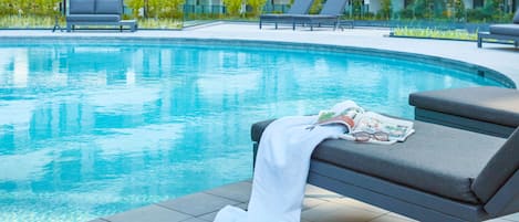 A modern hotel pool area with lounge chairs and a bench, surrounded by a well-maintained garden and a building with multiple balconies.