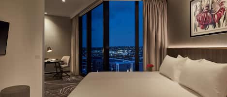 Habitación de hotel con una cama grande, un escritorio, una silla, una mesita y vistas a la ciudad de noche.