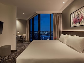 A hotel room with a large bed, a desk, a chair, a small table, and a view of the city at night.