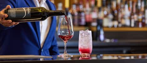A person pouring wine into a glass at a bar with a variety of bottles in the background.