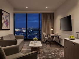 A modern hotel room with a large window offering a city view, a sofa, a small table with a vase, and a flat-screen TV mounted on the wall.