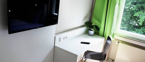 Et moderne værelse med et fladskærms-tv på væggen, et lille bord med en potteplante, en stol og et vindue med gardiner.