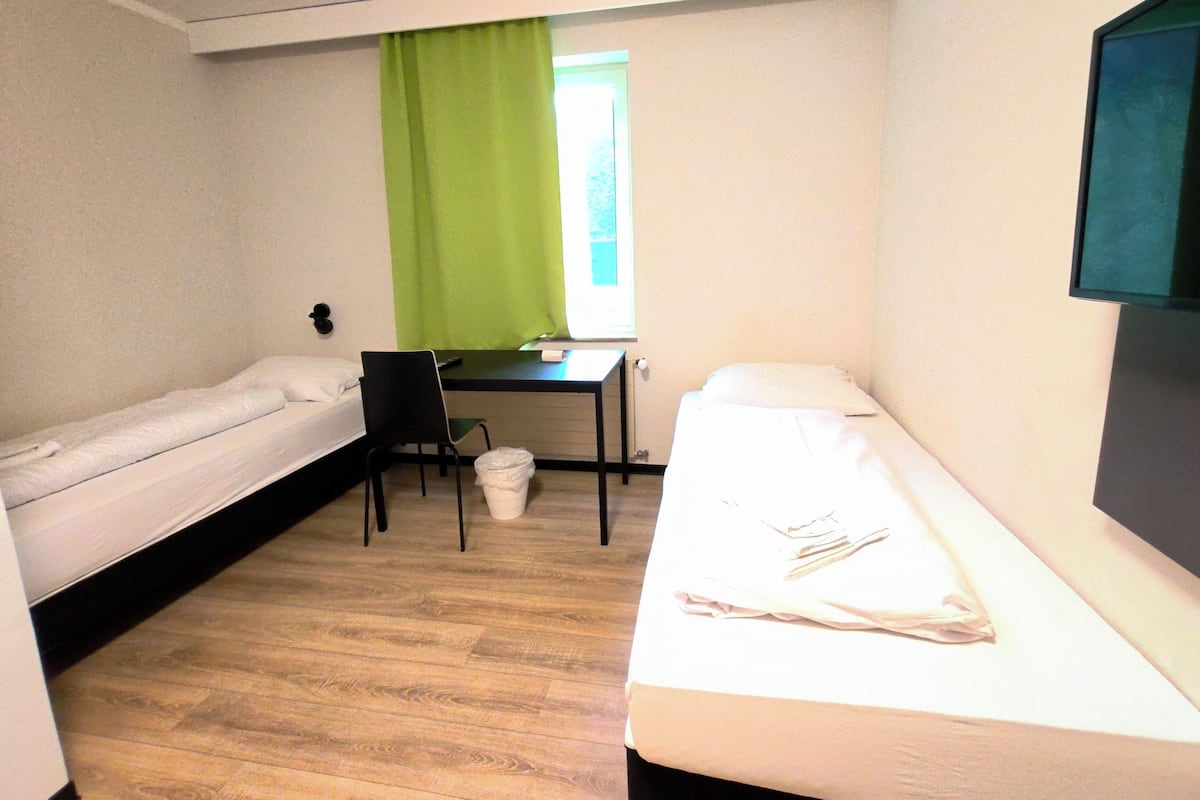 Ein kleines Zimmer mit zwei Einzelbetten, einem Schreibtisch, einem Stuhl und einem Fenster mit grünen Vorhängen.