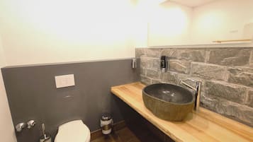Une salle de bain moderne avec un mur en pierre, un meuble-lavabo en bois, un grand lavabo, des toilettes et un bidet.