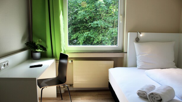 A hotel room with a bed, a desk, a chair, a potted plant, and a window with a view of trees. - Bendorf Hotel by Belvilla (Bendorf)