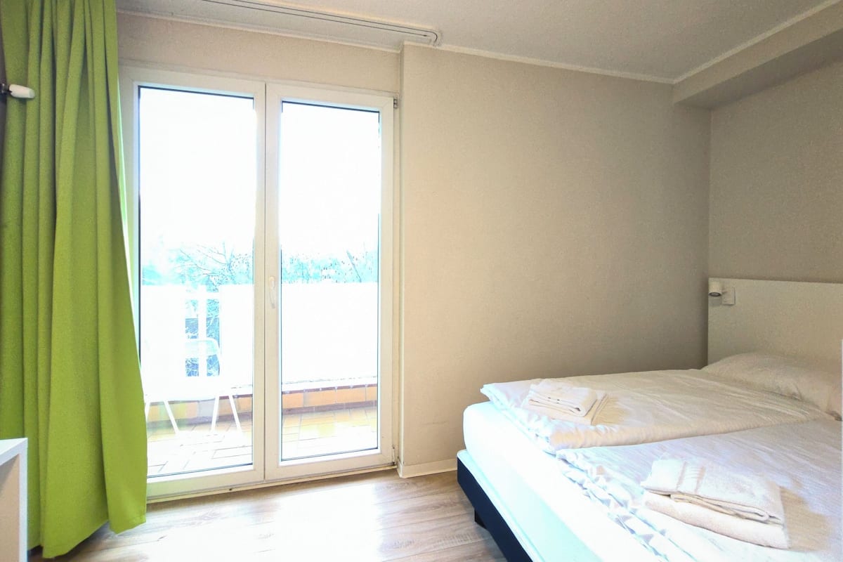 Ein Schlafzimmer mit Bett, einem Fenster mit Vorhängen und Blick ins Freie.