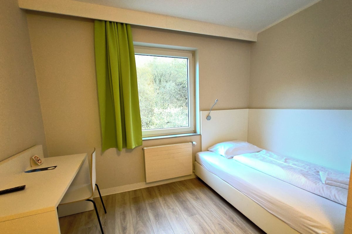 Ein kleines Hotelzimmer mit einem Bett, einem Schreibtisch, einem Stuhl, einem Fenster mit Blick auf Bäume und einer Heizung.