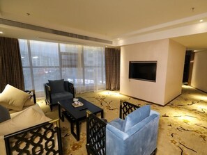 Habitación de hotel con sofá, sillones, una mesa de centro, televisor de pantalla plana y un amplio ventanal con cortinas.