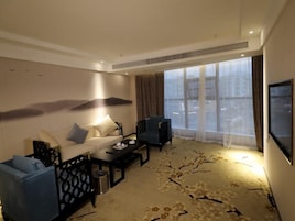 A hotel room with a sofa, armchairs, a coffee table, a TV, and a large window with curtains.