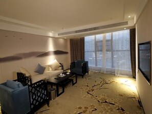 Une chambre d’hôtel avec un canapé, des fauteuils, une table basse, une télévision et une grande fenêtre avec des rideaux.