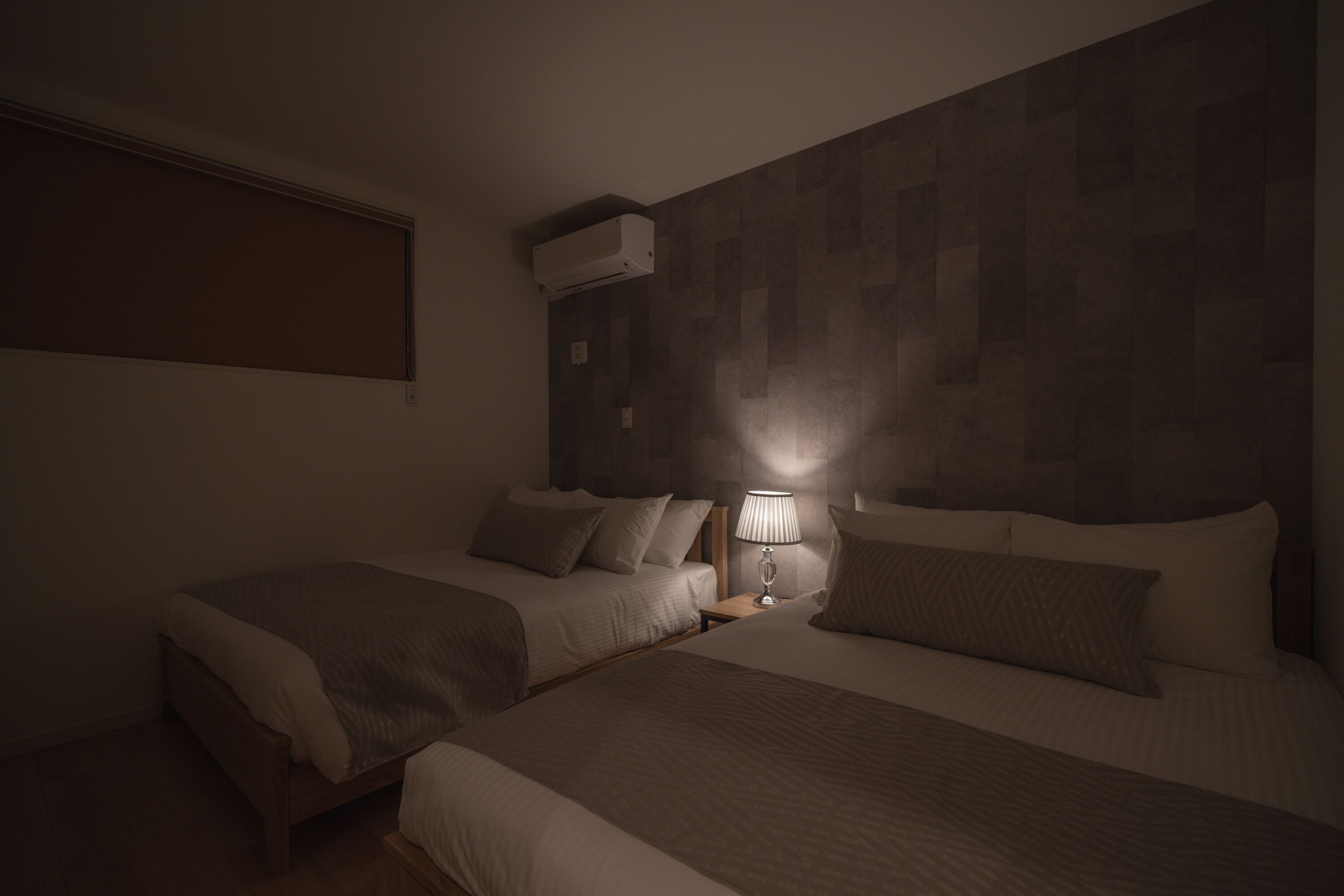 A hotel room with two beds, a nightstand with a lamp, and an air conditioning unit on the wall.