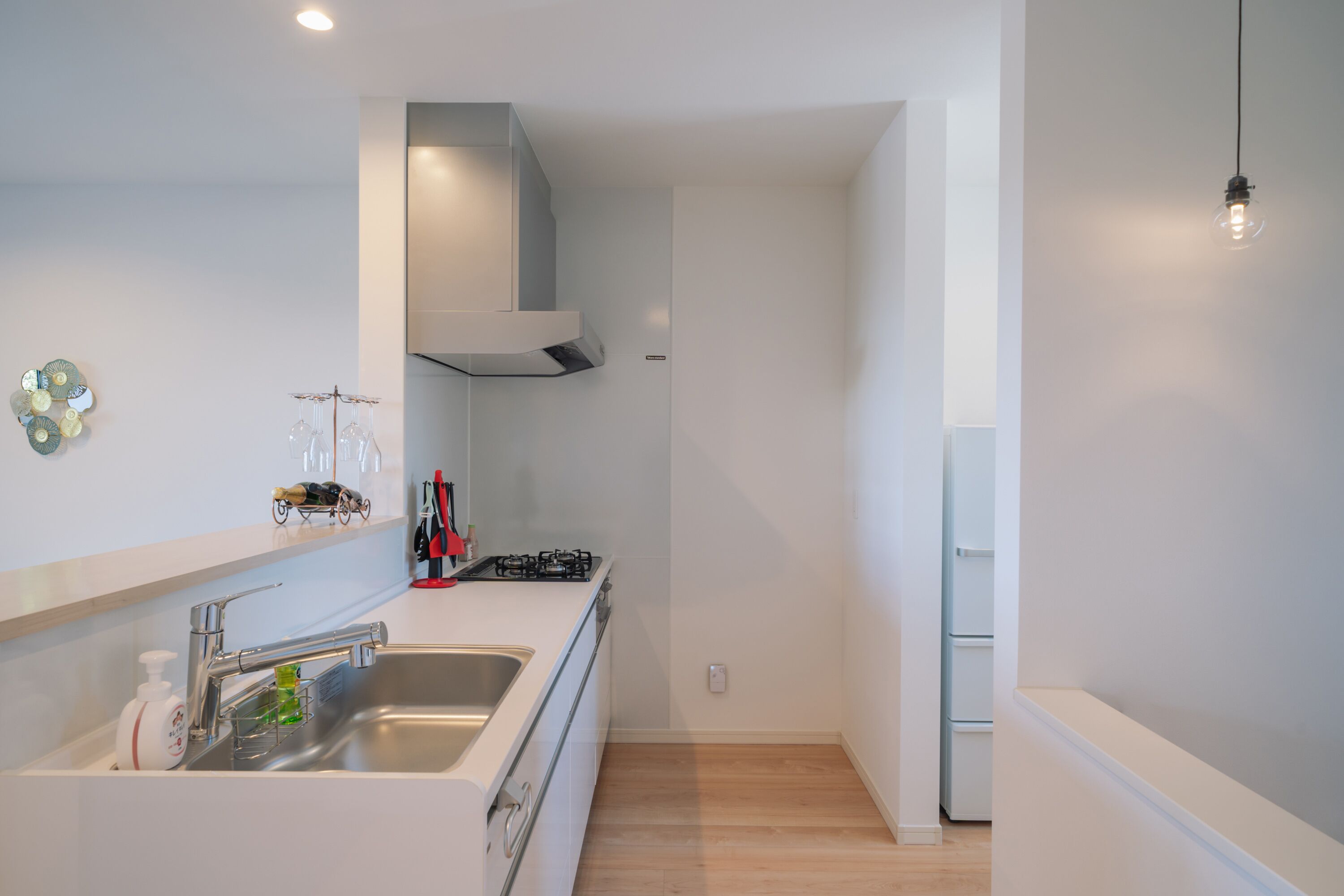 Une cuisine moderne dotée d’un évier en acier inoxydable, d’une cuisinière à gaz et d’un four encastré. On y trouve également un réfrigérateur blanc et un luminaire fixé au mur.