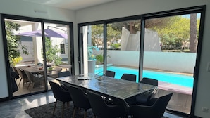 A modern dining area with a glass table and chairs, a black rug, and a view of a swimming pool and outdoor seating.