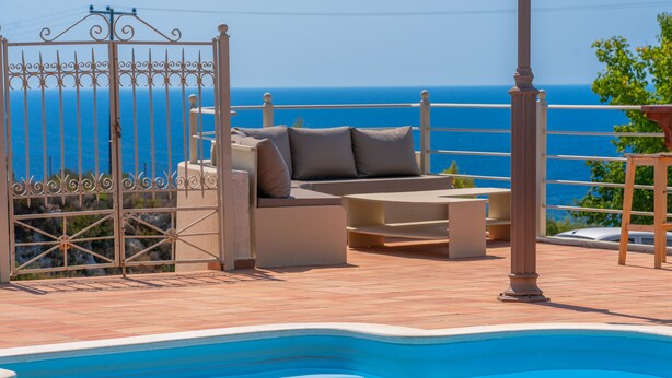 Un área junto a la piscina con sofá y mesa, una reja y vista al mar.