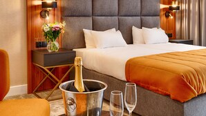 Hotellihuone, jossa on suuri sänky, yöpöytä maljakko täynnä kukkia, pöytä, jossa on samppanjapullo ja lasit, sekä moderni kattovalaisin.