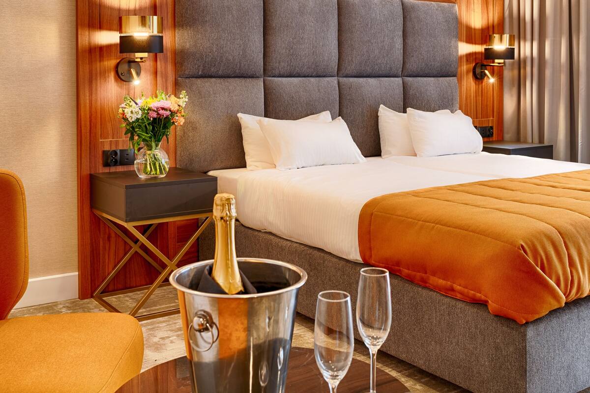 Ein Hotelzimmer mit einem großen Bett, einem Nachttisch mit Blumenvase, einem Tisch mit einer Champagnerflasche und Gläsern sowie einer modernen Deckenleuchte.
