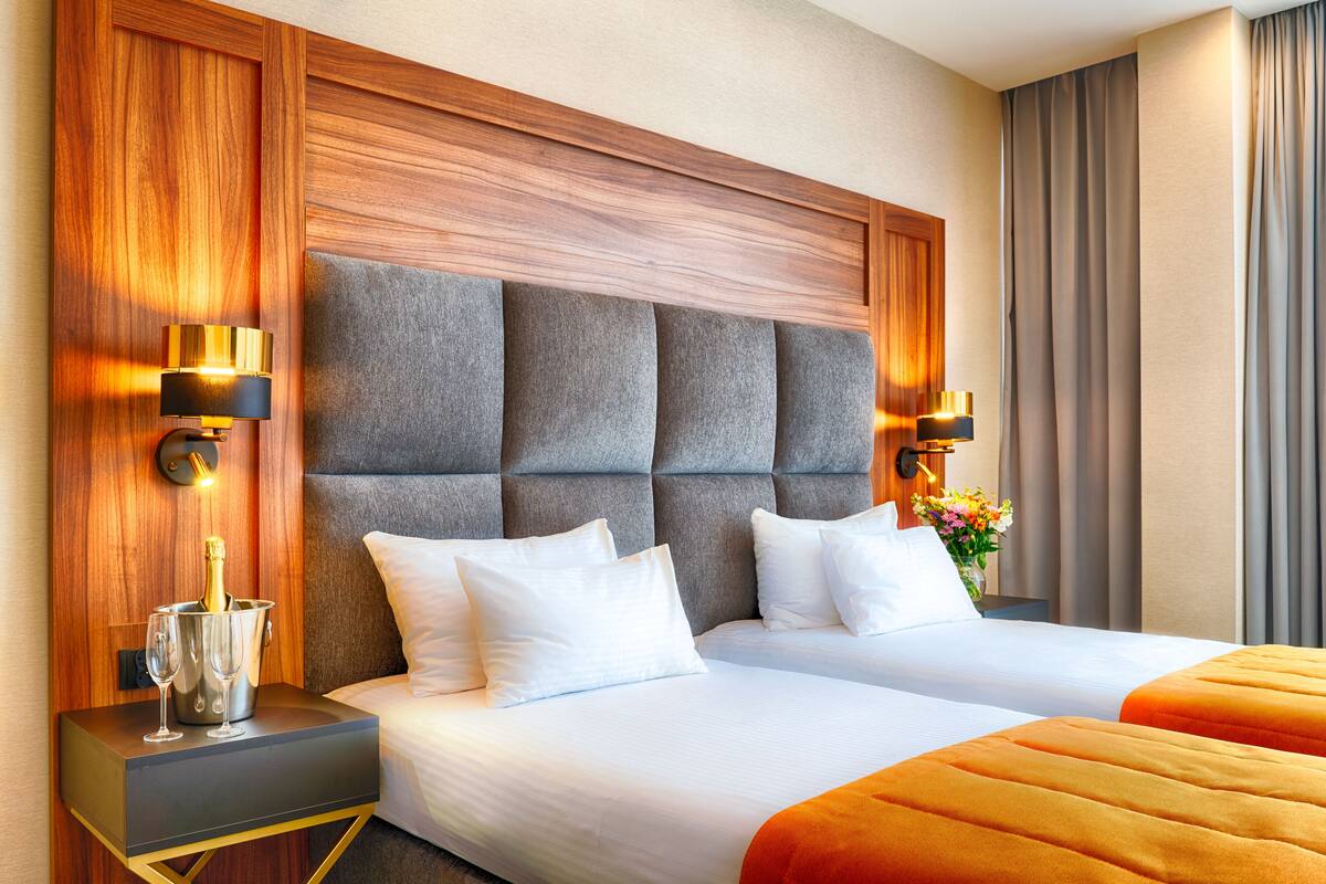 Ein Hotelzimmer mit zwei Betten, einem Holzkopfteil, Nachttischlampen, einem Nachttisch mit Champagnerkühlbox und einer Vase mit Blumen.