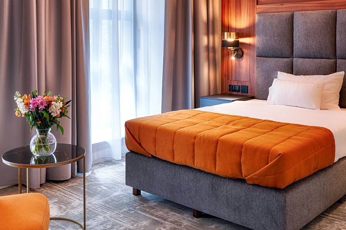 Ein modernes Hotelzimmer mit einem großen Bett, einem Nachttisch, einer Blumenvase und Blick ins Freie durch transparente Vorhänge.