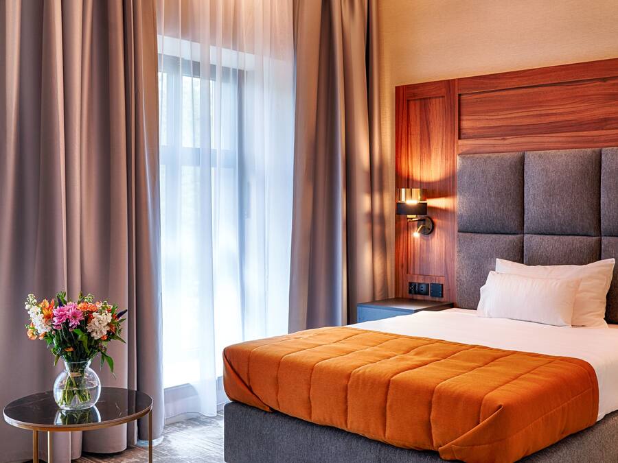 Moderni hotellihuone, jossa on suuri sänky, yöpöytä, maljakko täynnä kukkia ja näkymä ulos ohuiden verhojen läpi.