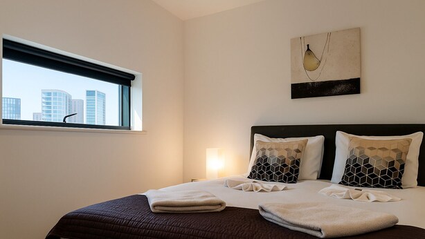 A hotel room with a large bed, two decorative pillows, a window with a city view, and a wall-mounted artwork.
