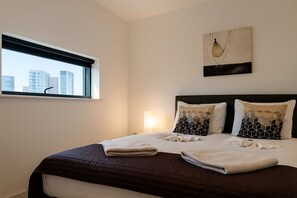 Habitación de hotel con una cama grande, dos almohadones decorativos, una ventana con vista a la ciudad y una obra de arte colgada en la pared.