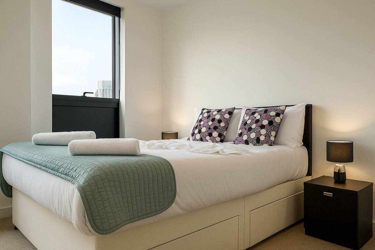 A modern hotel room with a large bed, a window with a city view, a bedside table with a lamp, and a nightstand.