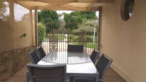 Un porche cubierto con una mesa y sillas, un muro de piedra y vistas a un jardín con una puerta metálica.
