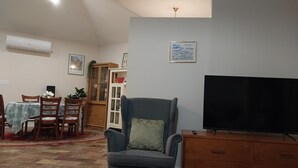 Una sala de estar con un sillón azul, una mesa de centro de madera, un televisor sobre un mueble de madera, un área de comedor con mesa y sillas, y un aparador con puertas de vidrio.