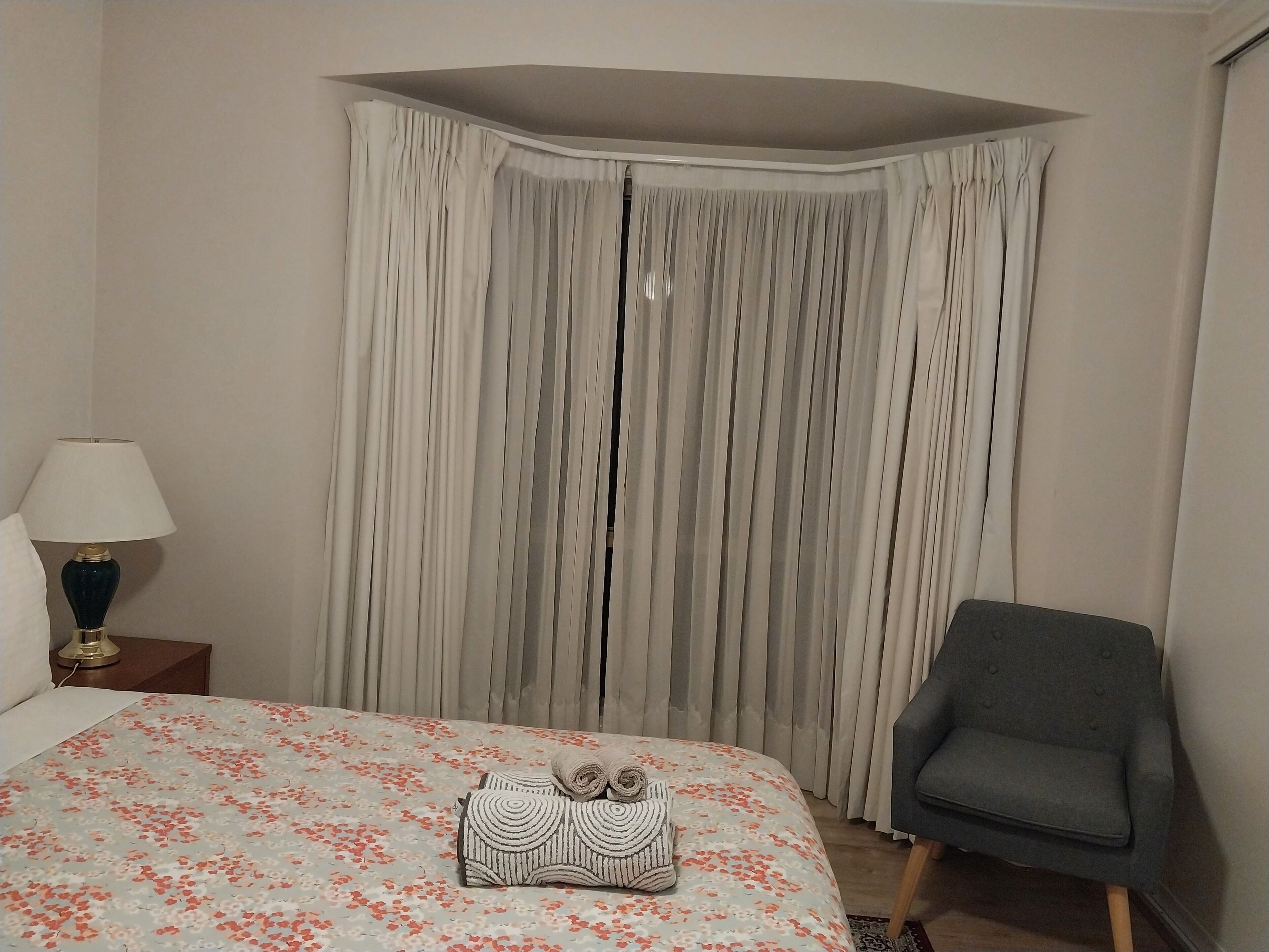 Ett hotellrum med en säng, en stol, en lampa och ett stort fönster med gardiner.