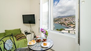 A room with a bed, a TV mounted on the wall, a table with pastries, and a view of a city and water through a window. - Apartment Luka (Dubrovnik)